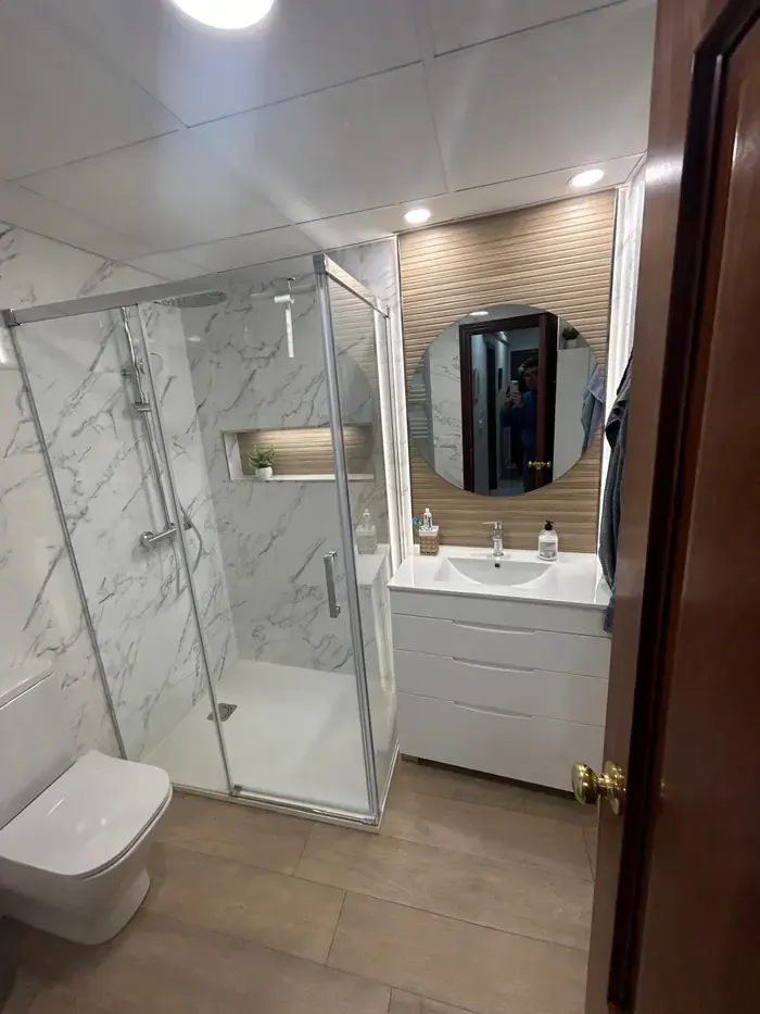 Baño con mampara, pared de mármol blanco y espejo redondo