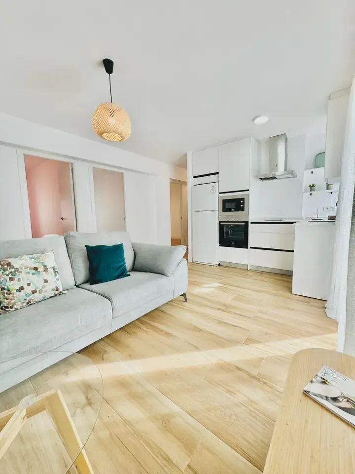Salón con sofá gris y suelo de madera clara, abierto a una cocina blanca