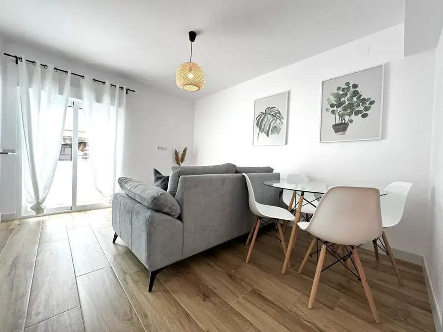 Salón comedor con sofá gris, mesa redonda y sillas blancas junto a un ventanal